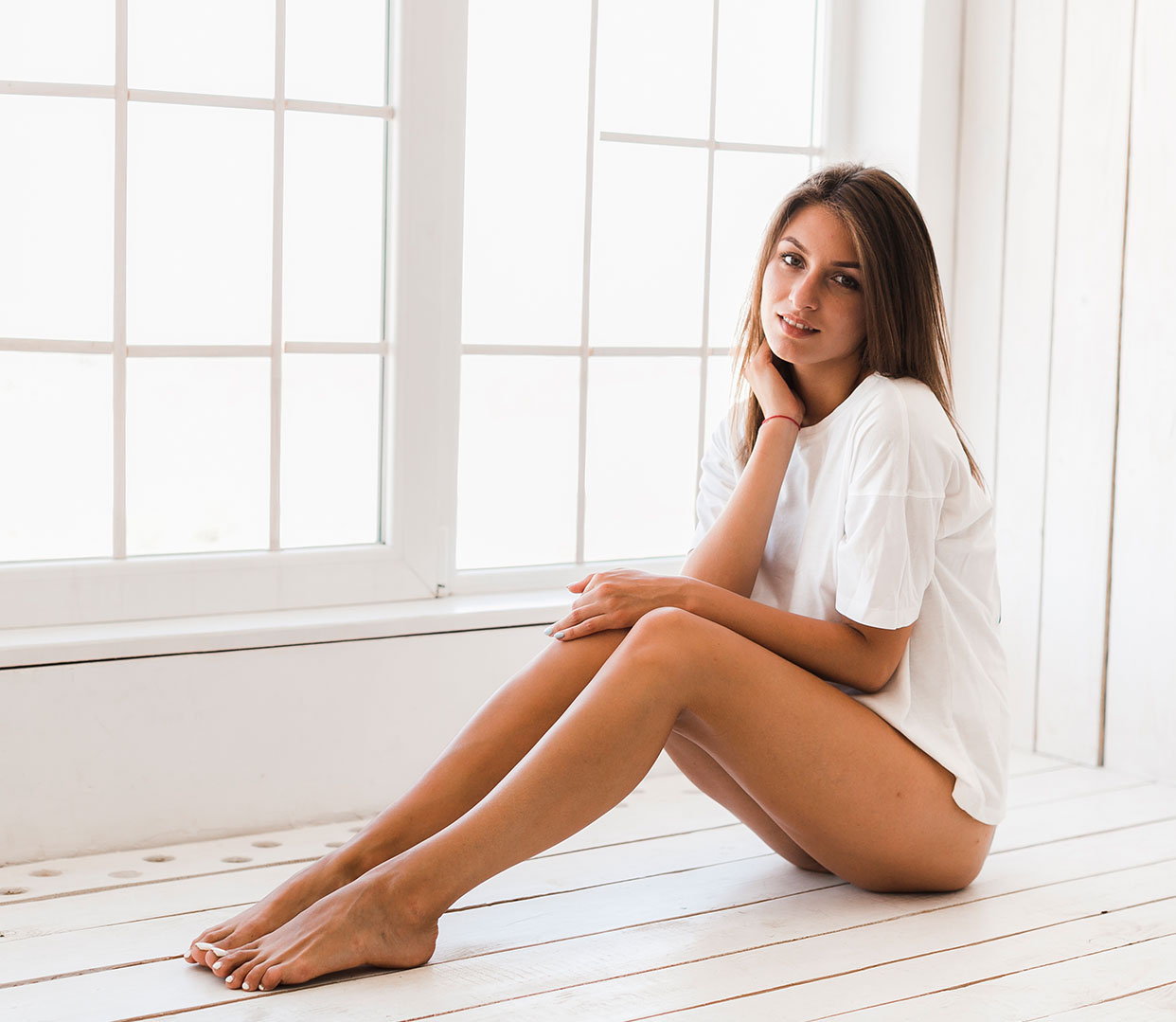 Une femme assise sur le sol, vêtue d'un t-shirt blanc, pose pour une photo. Elle a les jambes croisées et les mains posées sur les genoux. L'arrière-plan montre une fenêtre avec des moulures blanches et un sol en bois clair.